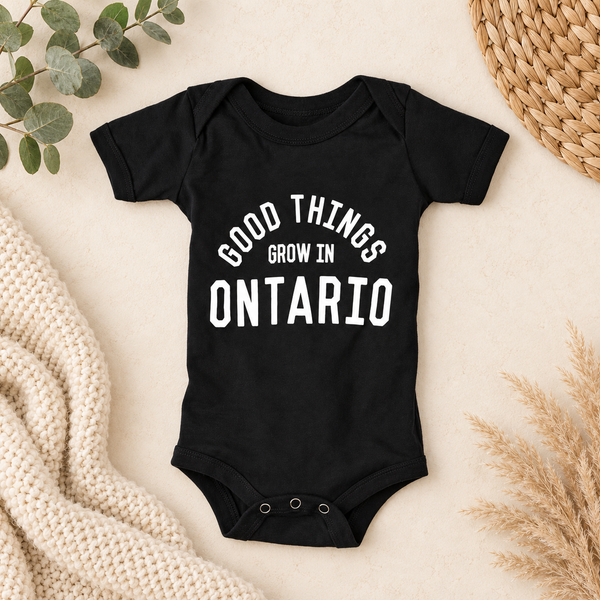 A black short-sleeve baby bodysuit with white text reading 'GOOD THINGS GROW IN ONTARIO' laid flat on a light beige surface with decorative knit fabric, dried grass, eucalyptus leaves, and a woven mat around it.