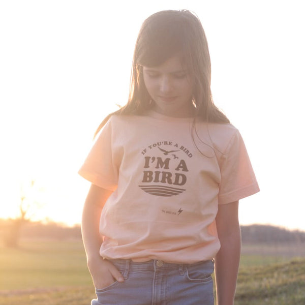 A young girl wearing a light peach-pink graphic t-shirt that reads 'IF YOU'RE A BIRD I'M A BIRD' in black lettering, paired with blue jeans, standing outdoors in a field during golden hour.